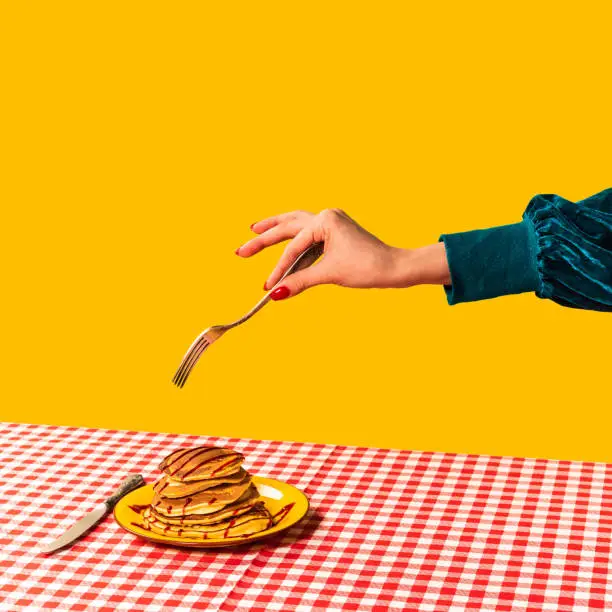 Photo of Food pop art photography. Female hand and sweet pancakes on plaid tablecloth isolated on bright yellow background. Vintage, retro style