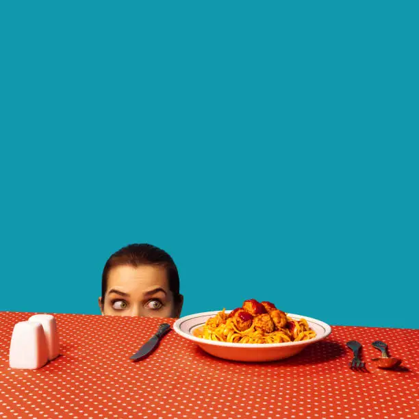 Photo of Young girl spying on spaghetti with meatballs on plaid tablecloth isolated on bright blue background. Food pop art photography. Vintage, retro style interior