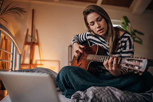 Beautiful woman musician playing acoustic guitar composing a song on bed in bedroom at home.