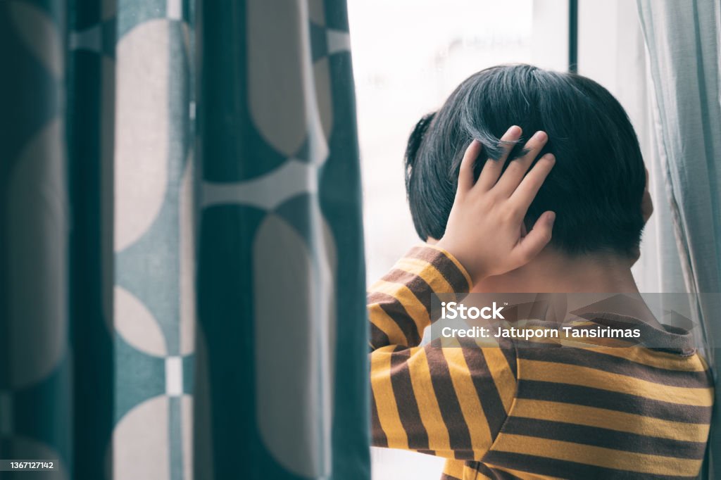Little Asian boy stand by window, hand cover ears, face down, crying and screaming. Little Asian boy wear long sleeve stripe shirt stand by window, hand cover ears, crying and screaming. Feeling frustration, anxiety, fear, stressed. Domestic violence, Autism, Bipolar disorder and mental illness concept. Autism Stock Photo