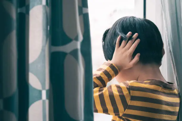 Little Asian boy wear long sleeve stripe shirt stand by window, hand cover ears, crying and screaming. Feeling frustration, anxiety, fear, stressed. Domestic violence, Autism, Bipolar disorder and mental illness concept.