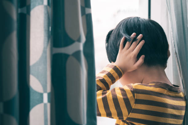 mały azjatycki chłopiec stoi przy oknie, zakrywa uszy, twarzą w dół, płacze i krzyczy. - bullying color image people photography zdjęcia i obrazy z banku zdjęć