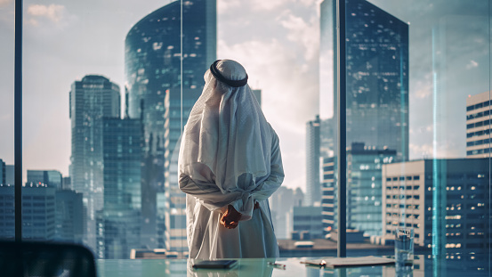 Exitoso hombre de negocios musulmán con atuendo blanco tradicional de pie en su oficina moderna mirando por la ventana a la gran ciudad con rascacielos. Exitoso concepto de empresario saudí, emiratí y árabe. photo