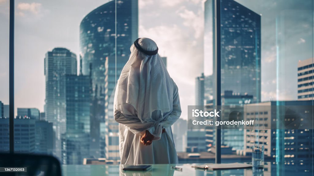 Successful Muslim Businessman in Traditional White Outfit Standing in His Modern Office Looking out of the Window on Big City with Skyscrapers. Successful Saudi, Emirati, Arab Businessman Concept. Saudi Arabia Stock Photo