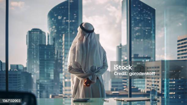 Successful Muslim Businessman In Traditional White Outfit Standing In His Modern Office Looking Out Of The Window On Big City With Skyscrapers Successful Saudi Emirati Arab Businessman Concept Stock Photo - Download Image Now