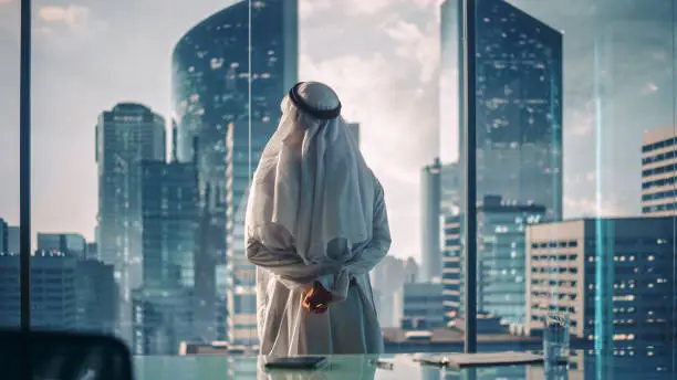 Photo of Successful Muslim Businessman in Traditional White Outfit Standing in His Modern Office Looking out of the Window on Big City with Skyscrapers. Successful Saudi, Emirati, Arab Businessman Concept.