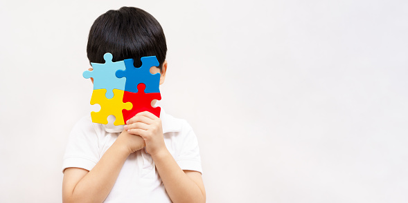 Studio shot of Asian young small child wear white shirt holding colorful jigsaw puzzle up cover his face in white background with copy space for autism spectrum and mental health disability symbol for World Autism Awareness day concept.