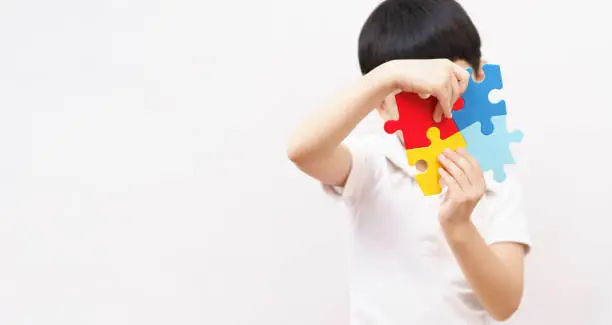 Studio shot of Asian young small child wear white shirt holding colorful jigsaw puzzle up cover his face in white background with copy space for autism spectrum and mental health disability symbol for World Autism Awareness day concept.