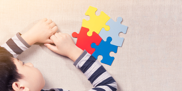 Studio table top view of autistic Asian boy lie face on table, hand holding fingers with frustration and stress, sadness with colorful jigsaw puzzle on beige linen cloth on table. World autism awareness day support concept.