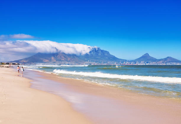 Table Mountain rises in the distance as two women stroll on the beach at Milnerton, Cape Town, South Africa Cape Town, South Africa - January 25, 2022: The world-famous landmark of Table Mountain, with the city of Cape Town nestled at its foot, rises above the sea in Table Bay as two women stroll along the sandy beach. The mountain is shrouded by a typical bank of cloud known to locals as the tablecloth. table mountain and the tablecloth stock pictures, royalty-free photos & images