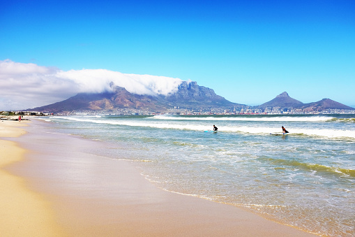 Cape Town, South Africa - January 25, 2022: The world-famous landmark of Table Mountain, with the city of Cape Town nestled at its foot, rises above the sea in Table Bay as surfers wearing wetsuits ride in on the waves. The mountain is shrouded by a typical bank of cloud known to locals as the tablecloth.