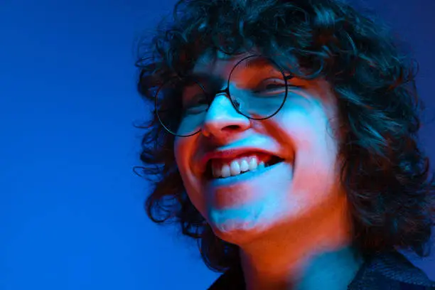 Photo of Portrait of young smiling man, student in black hoodie isolated on dark blue background in neon light. Concept of emotions, facial expression, youth, aspiration, sales