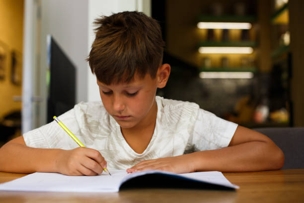 istock A young boy in a white T-shirt is sitting at the table. He is writing homework in his notebook. 1367137002