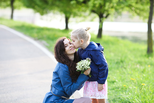 Little girl in blue jacket kissing her mother and giving her a bouquet of lilies of the valley on a sunny spring day. Mother's day concept. Selective focus. Soft focus.