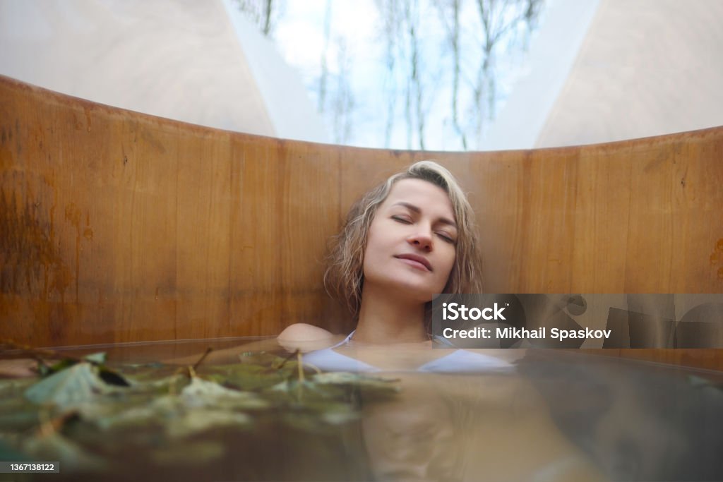 A young woman takes hot bath in oak barrel in the winter outside. Steam barrel and spa center. A young woman in white swimsuit takes hot bath in oak barrel in the winter outside. Steam barrel and spa center. Spa Stock Photo