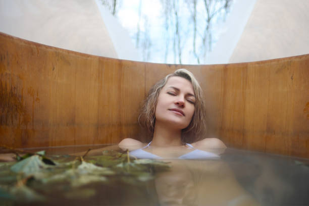 A young woman takes hot bath in oak barrel in the winter outside. Steam barrel and spa center. A young woman in white swimsuit takes hot bath in oak barrel in the winter outside. Steam barrel and spa center. thermal pool stock pictures, royalty-free photos & images
