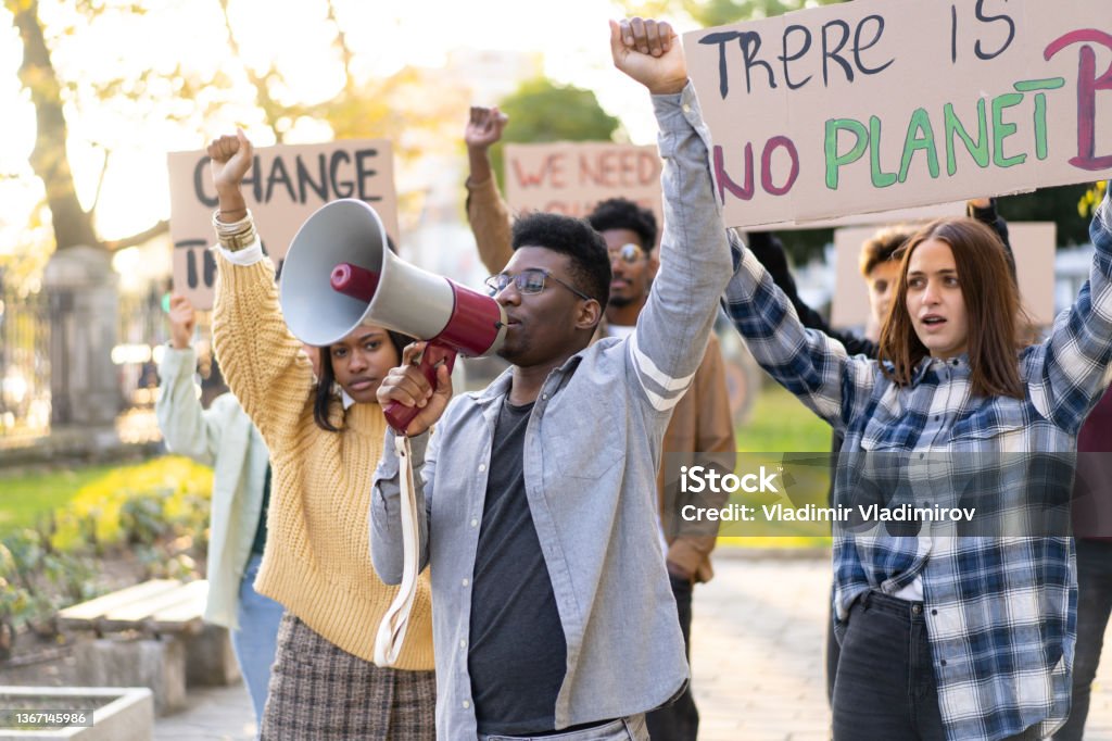 Students protesting against climate change A group of young people demonstrating in a local park, holding placards and with their arms raised in the air as an African man uses a megaphone to broadcast their message for environmental issues Fridays for Future Stock Photo