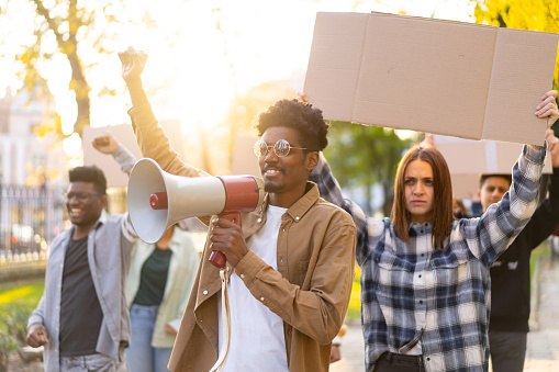 A group of activists holding blank banners in the air while their leader uses a megaphone, in a public park on a sunny day