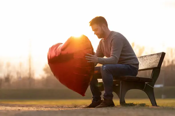 Sad young man holding up a heart-shaped balloon sitting on a bench in the park and looking at the balloon