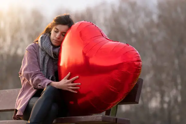 Photo of Sad young woman hugging a heart-shaped balloon sitting on a bench in the park