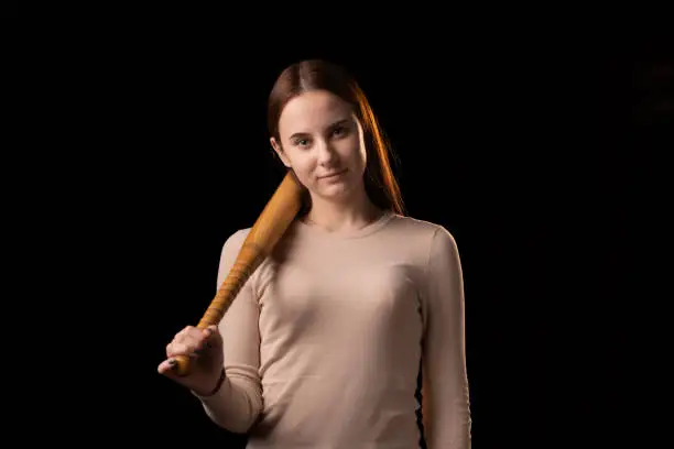 Photo of A young woman holds a bat in her hands and calmly looks into the camera. Black background. Self-defense, psychological protection, mental strength, inner peace concept.