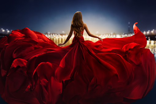 Mujer de moda en rojo revoloteando Vestido retrovisor lateral vista trasera. Modelo glamour bailando con tela de seda larga volando sobre el viento sobre el paisaje de luz de la ciudad del cielo nocturno photo