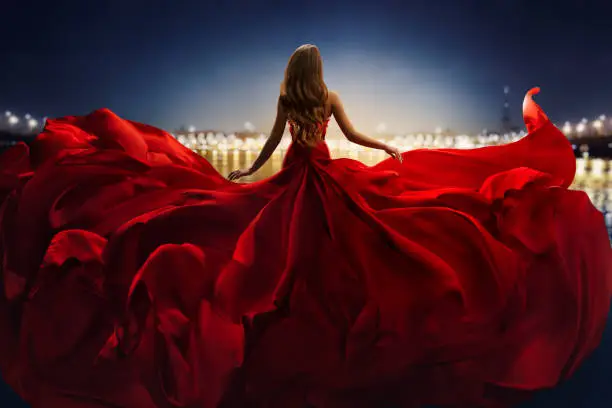 Fashion Woman in Red fluttering Dress Back Side Rear View. Glamour Model Girl dancing with Long Silk Fabric flying on Wind over Night Sky City Light Landscape