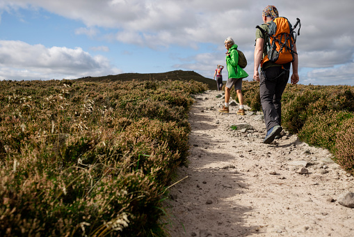 A woman wearing a backpack on a hike in Rothbury, Northumberland. She is being followed by her son while looking back at him. They are both walking along a footpath between shrub land.