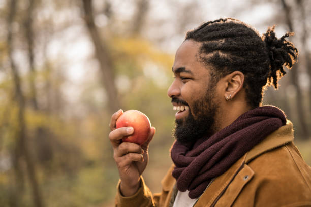 счастливый молодой афроамериканец ест яблоко во время прогулки на свежем воздухе на природе. - apple healthy eating eating black стоковые фото и изображения