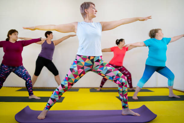 durante una clase de yoga, las mujeres están realizando la pose de guerrero - women yoga yoga class mature adult fotografías e imágenes de stock