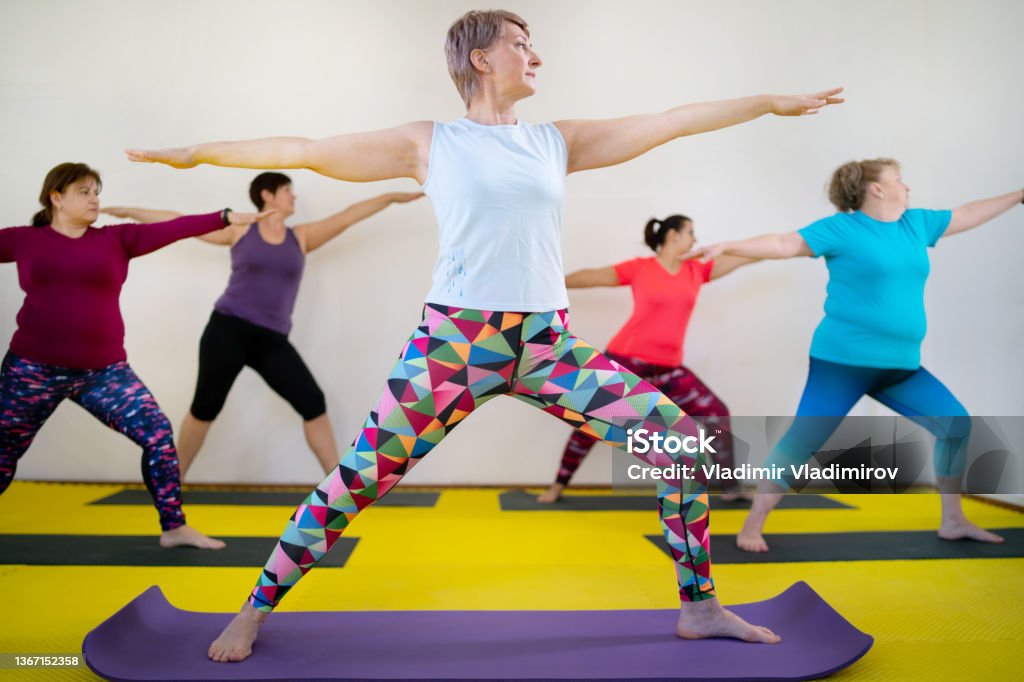 During a yoga class, ladies are performing the warrior pose A group of mid adult women are balancing with their legs wide apart as they face forward and hold their arms outstretched in a  yoga hall standing on exercise mats Obesity Stock Photo
