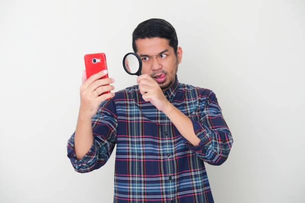 istock Adult Asian man looking to mobile phone using magnifying glass with suspicious expression 1367157414