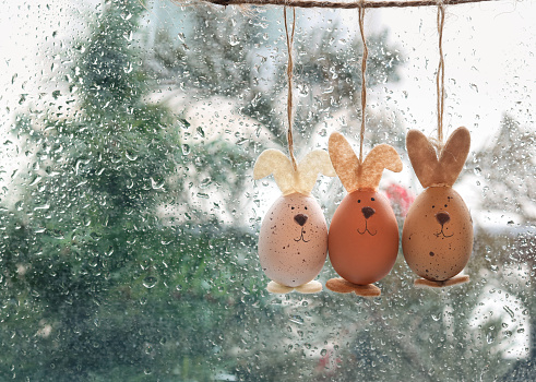 A rabbit Easter egg ornament hanging against the window glass with drop of the rain in the background, vintage atmosphere and copy space. A festive holiday decoration