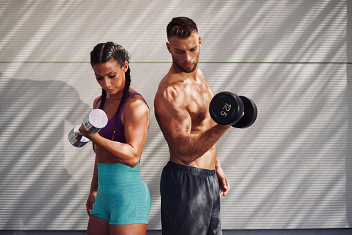 Athletic couple pumping up muscles with dumbbells outdoors