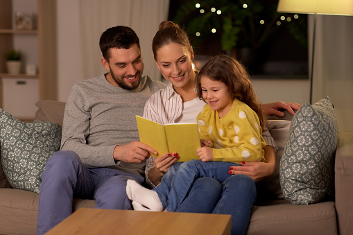 family, leisure and people concept - happy father, mother and little daughter reading book at home at night