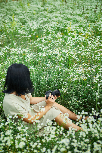 Female photographer sitting outdoors on flower field landscape holding a camera, woman hold digital camera in her hands. Travel nature photography, space for text, top view.