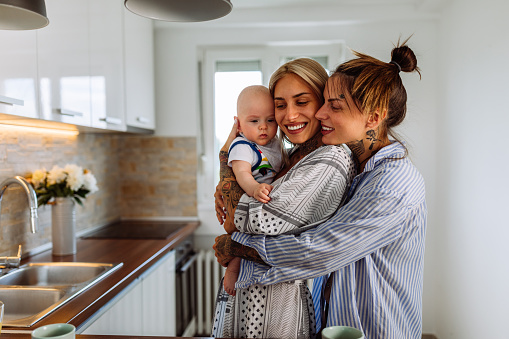 Happy young lesbians playing with baby boy while standing in the kitchen during morning