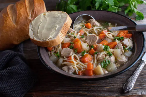 Fresh and homemade cooked chicken noodle soup served with baguette and butter in a rustic enamel bowl on wooden table.