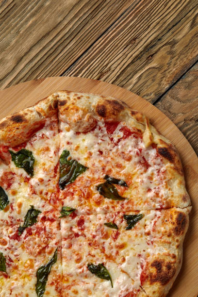 Uncut Italian pizza served with fresh basil leaf on Wooden board on dark texture background. Hot pizza with tomatoes and pesto sauce