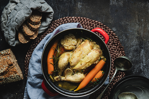 Top view of clear Chicken broth with carrots, onions, garlic, rosemary and thyme in a metal casserole with wholegrain bread on the side. Healthy chicken soup with organic vegetables in a pan with bread on kitchen counter.