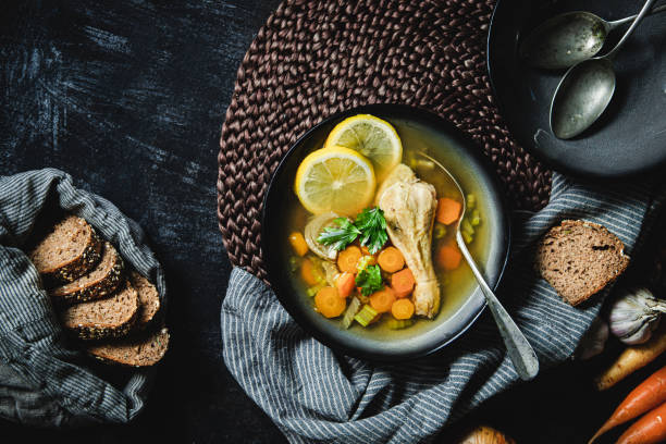 Top view of a bowl of chicken broth with vegetables and wholegrain bread on the kitchen counter. Healthy chicken soup served on table.