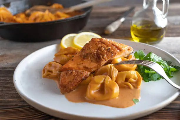 Delicious savory italian fish dish with roasted salmon fillet, cooked with tortellini in a delicious tomato cream sauce. Served on a plate with lemon on wooden table background. Ready to eat
