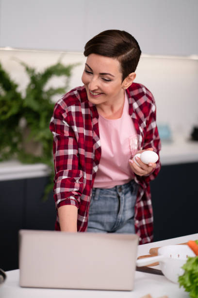 istock Woman in kitchen using laptop holding eggs in hand smiling while reading recipe or having a video call. Woman in red plaid shirt in modern kitchen interior 1367172244