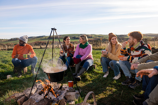 A group of volunteers wearing warm casual clothing and accessories on a sunny cold winters day. They are resting and having a lunch break from working on a community farm, planting trees and performing other sustainable and environmentally friendly tasks. They are gathered round a campfire which they are cooking soup over in a pot, talking and enjoying the hot soup.