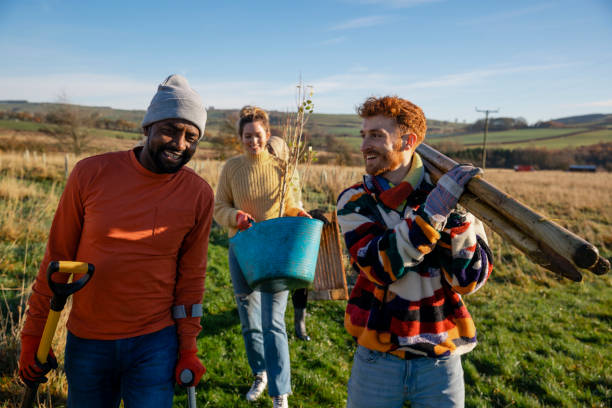 Helping Others A multiracial group of people wearing warm casual clothing on a sunny cold winters day. Carrying posts and a young tree, friends walk with a man using a crutch. They are working on a community farm, planting trees and performing other sustainable and environmentally friendly tasks. walking for a cause stock pictures, royalty-free photos & images