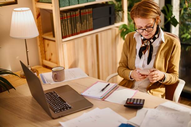 istock Happy senior woman using mobile phone while working at home with laptop 1367180264