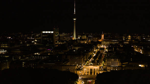 A high-flying drone shot of the Brandenburg Gate at night. In the background the illuminated Red Town Hall and the Berlin TV Tower.