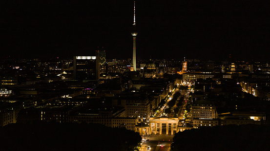 A high-flying drone shot of the Brandenburg Gate at night. In the background the illuminated Red Town Hall and the Berlin TV Tower.