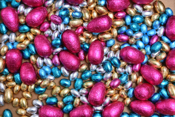 istock Small pink, blue, silver and gold foil wrapped chocolate easter eggs, with large pink eggs against a pale wood background. 1367188702