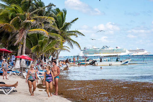 Mahahual, Mexico - January 6, 2022: A large number of tourists, most of them cruise passengers, on the beach in Mahahual. The cruise ships in the background include the Carnival Pride and Liberty of the Seas.